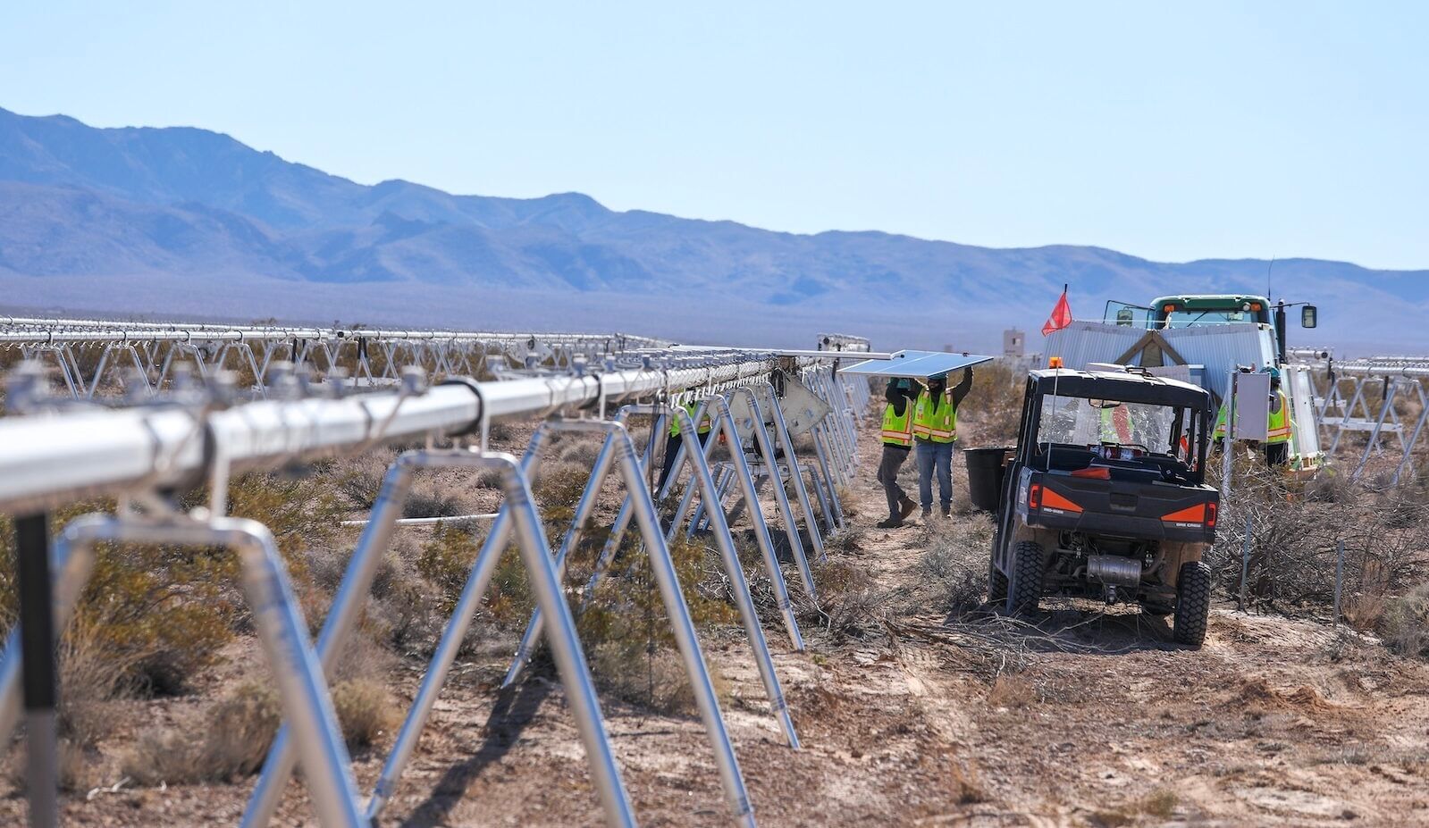 Two workers carrying a panel to attach to a solar grid in a desert landscape surrounded by moutntains