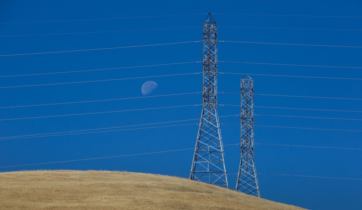 Two transmission towers on dry-grass hilll with blue sky and moon visible behind