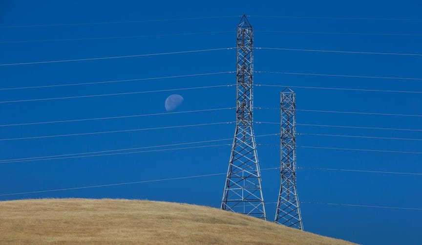 Two transmission towers on dry-grass hilll with blue sky and moon visible behind