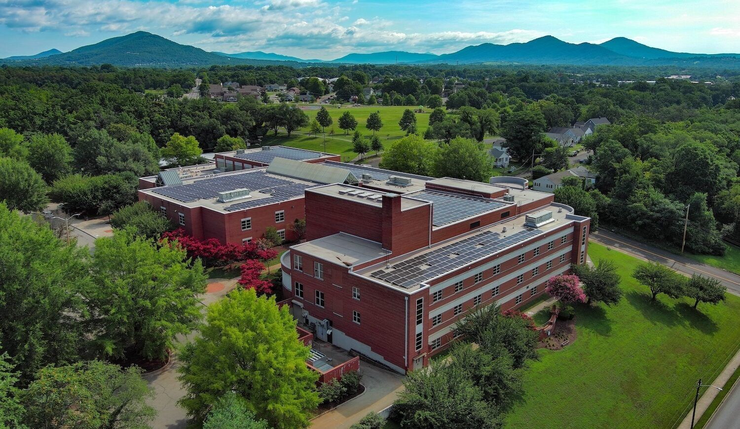 Overhead view of red school building with solar panels on its roof, surrounded by lawn and trees with mountains in distance