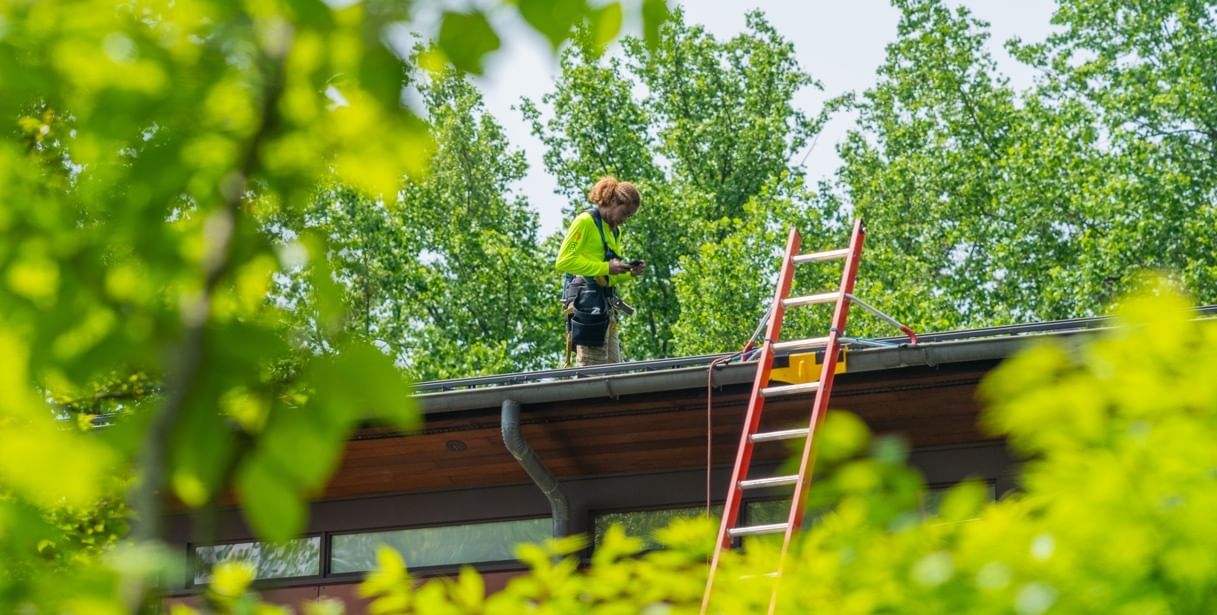 A person standing on roof looking at a smartphone wearing a lime-green top and tan pants. Green trees surround the roof