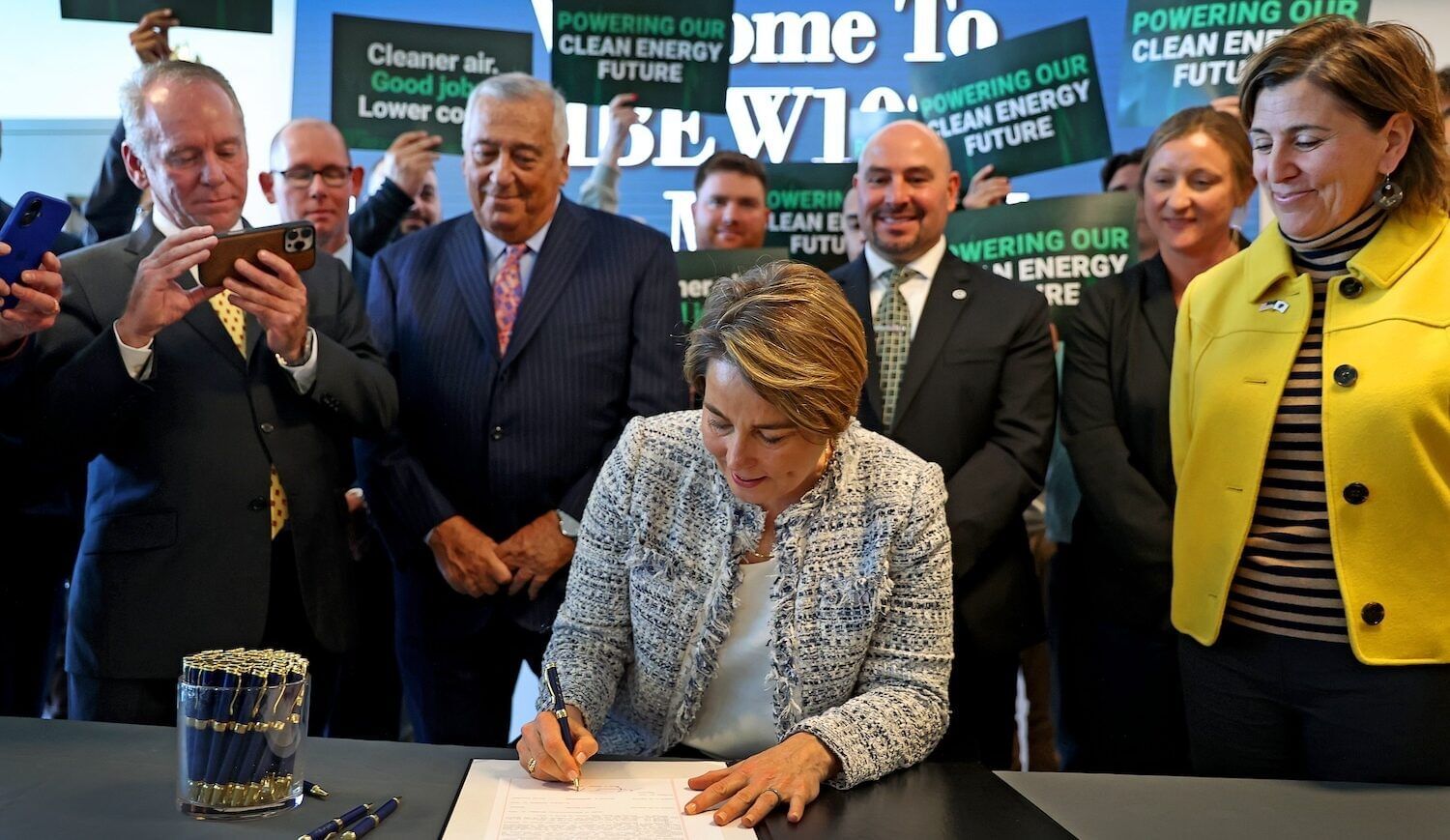 Woman sitting and signing a paper, surrounded by people holding signs reading "Powering our clean energy future"
