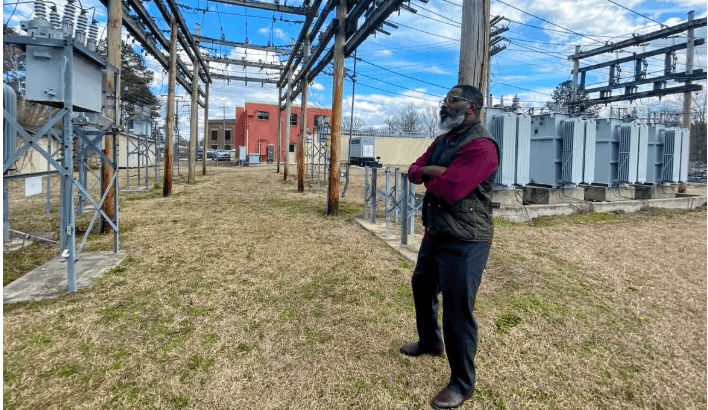 A man in dark pants and vest, maroon long-sleeve top, stands outside on dry grass under power lines near electric equipment