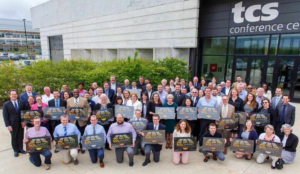 A group of people in business casual clothes posing in front of a conference center with signs that say "SolSmart"