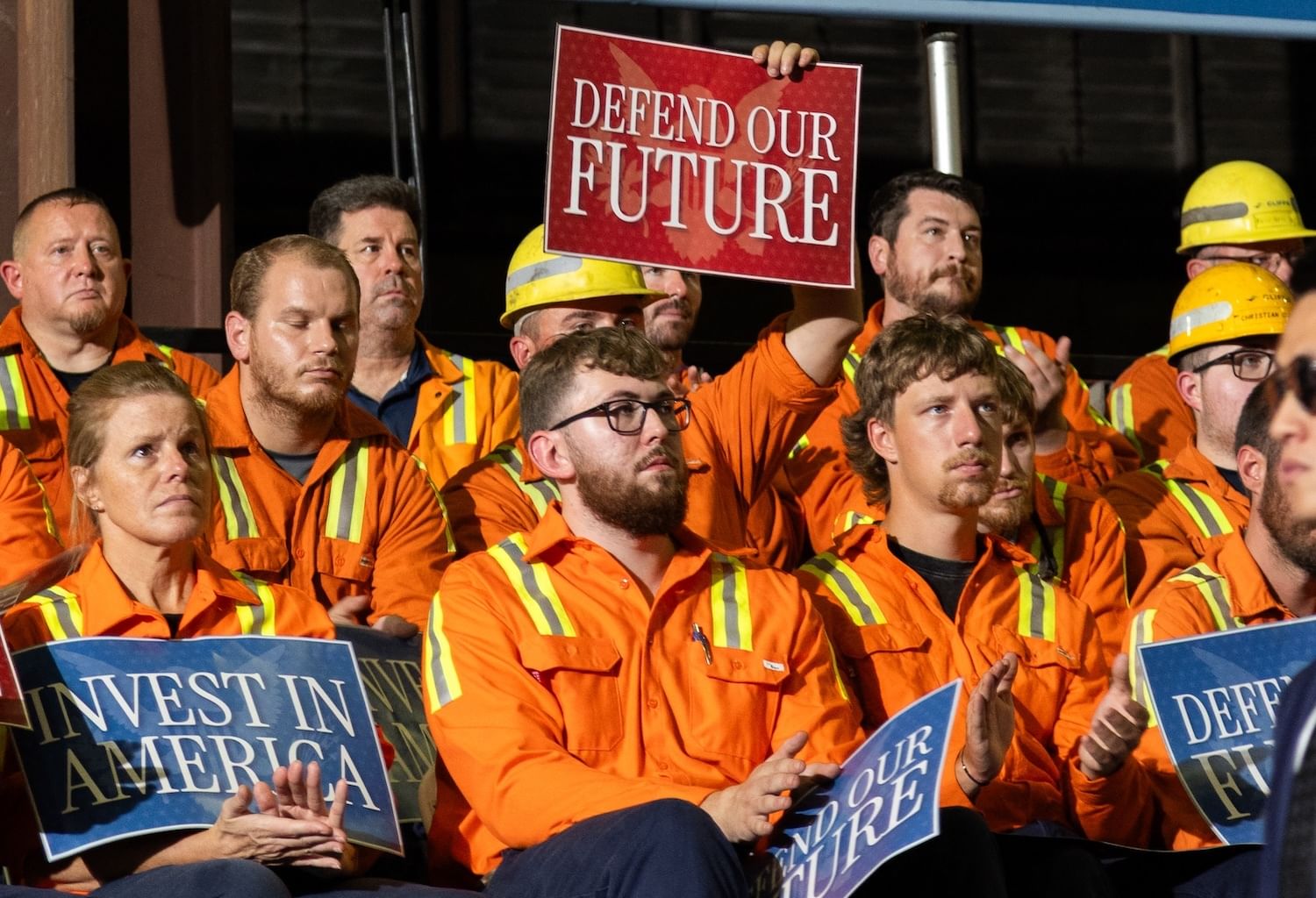 Seated workers in orange safety shirts holding "Invest in America" signs