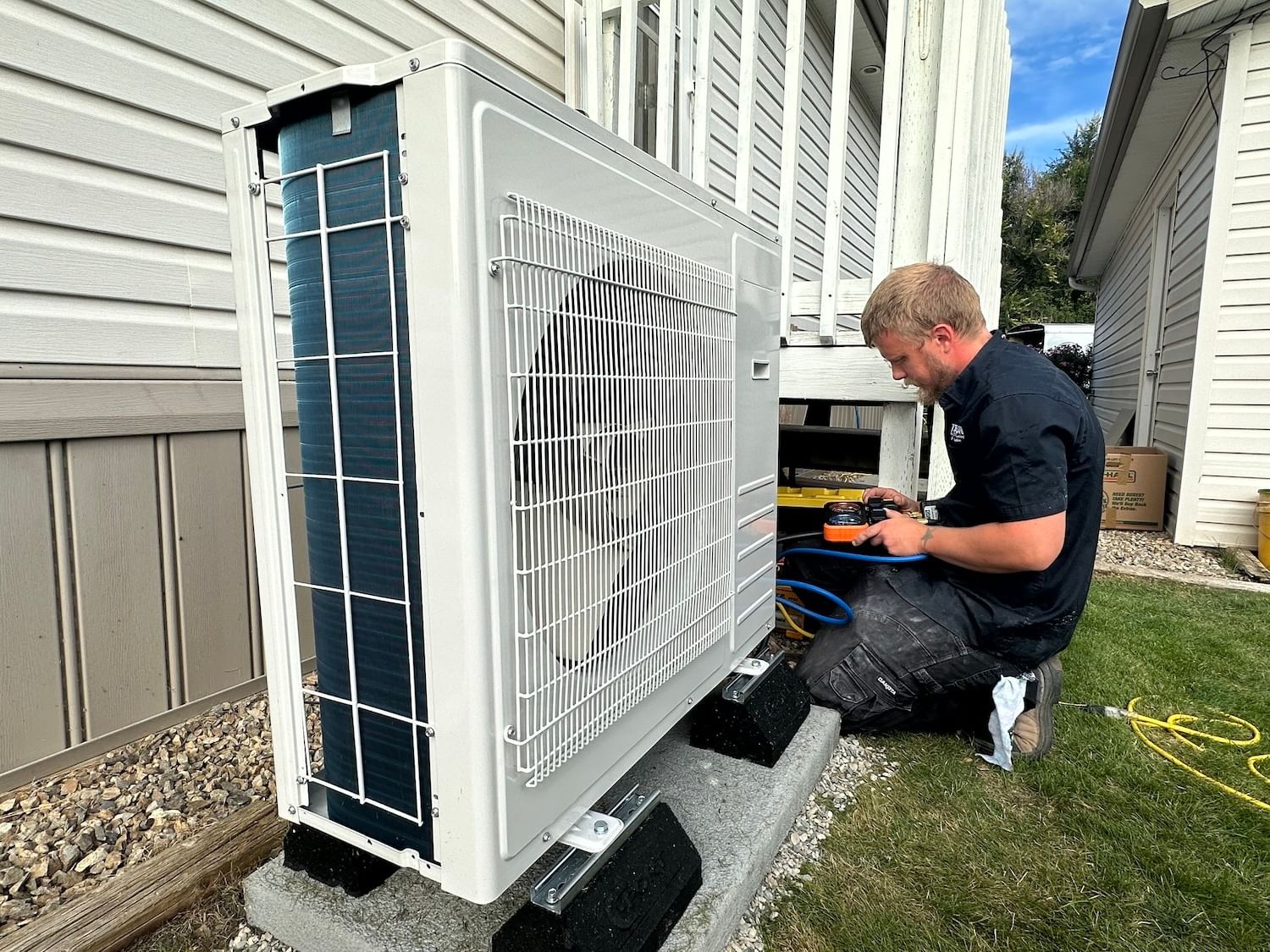 A man kneels on a lawn next to a heat pump buy the side of a house