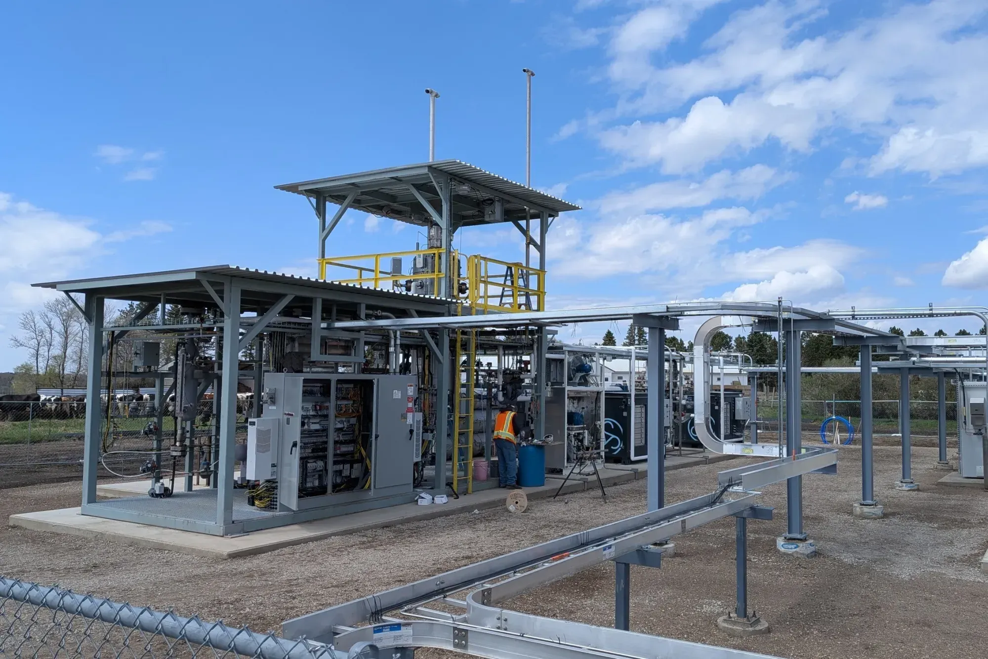 Gray tanks, pipes, and sheds on a dirt plot; one orange-vested worker in the center