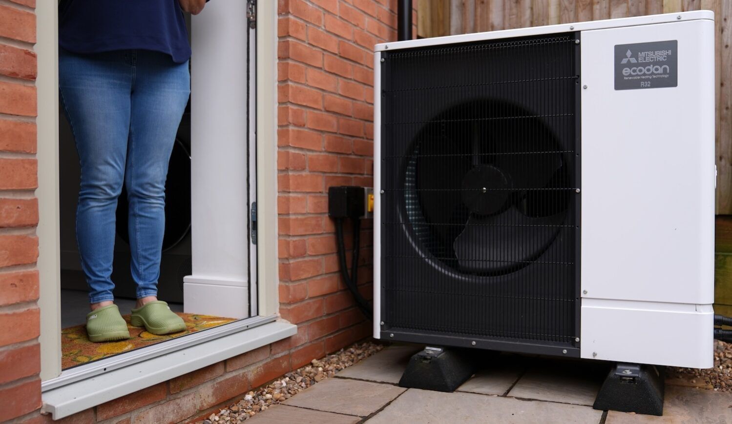 A  Mitsubishi Electric Ecodan R32 air source heat pump by a home's open doorway, in which a woman's legs in jeans are seen