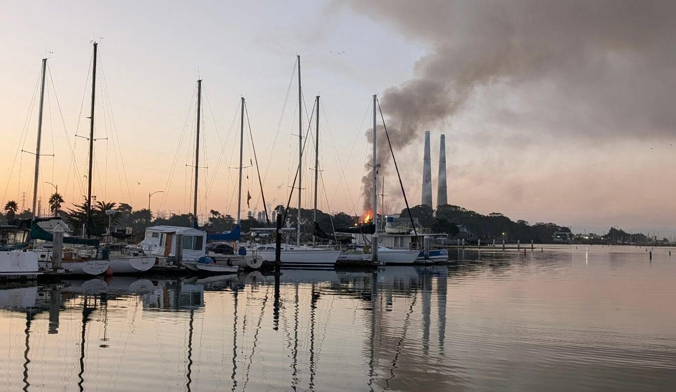 A view of a harbor with boats with flames in the background next to two smokestacks