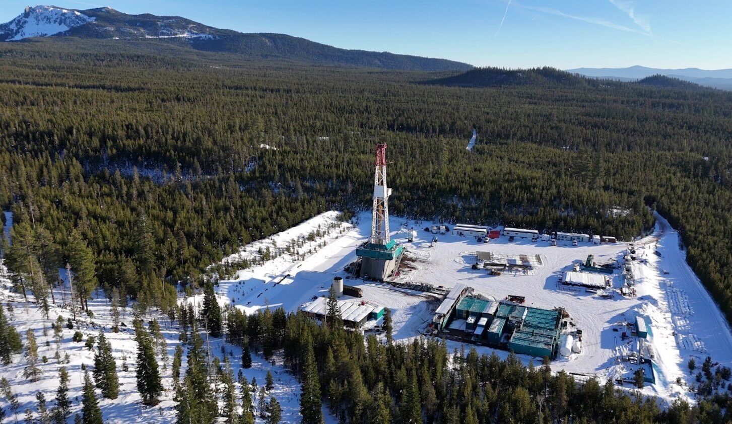 Overview of a snow-covered geothermal site surrounded by tall trees with mountain in background