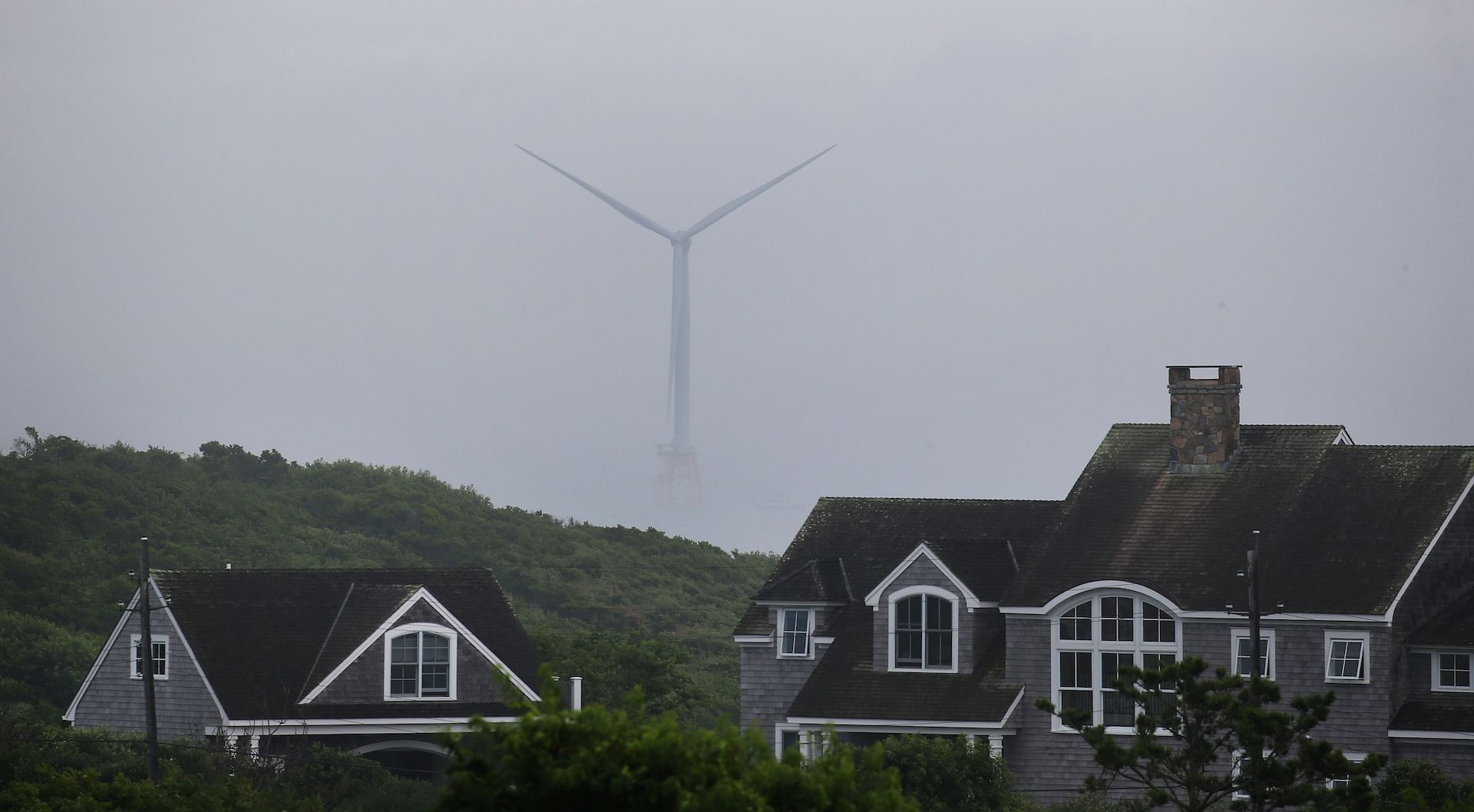 One wind turbine enshrouded by fog stands above two buildings and treetops