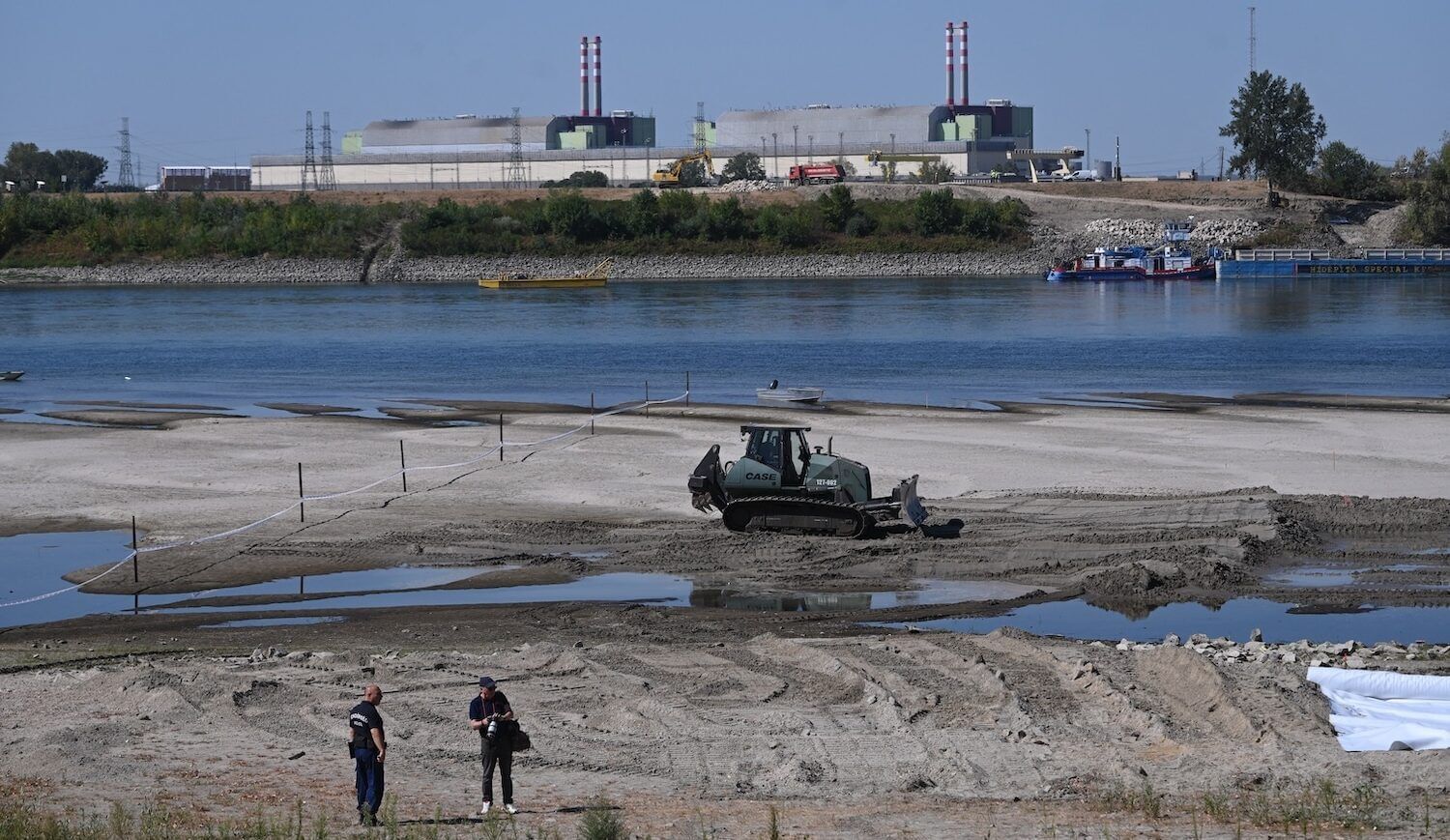 Two workers and construction equipment, low river, and nuclear plant in distance