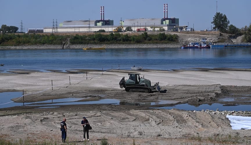 Two workers and construction equipment, low river, and nuclear plant in distance