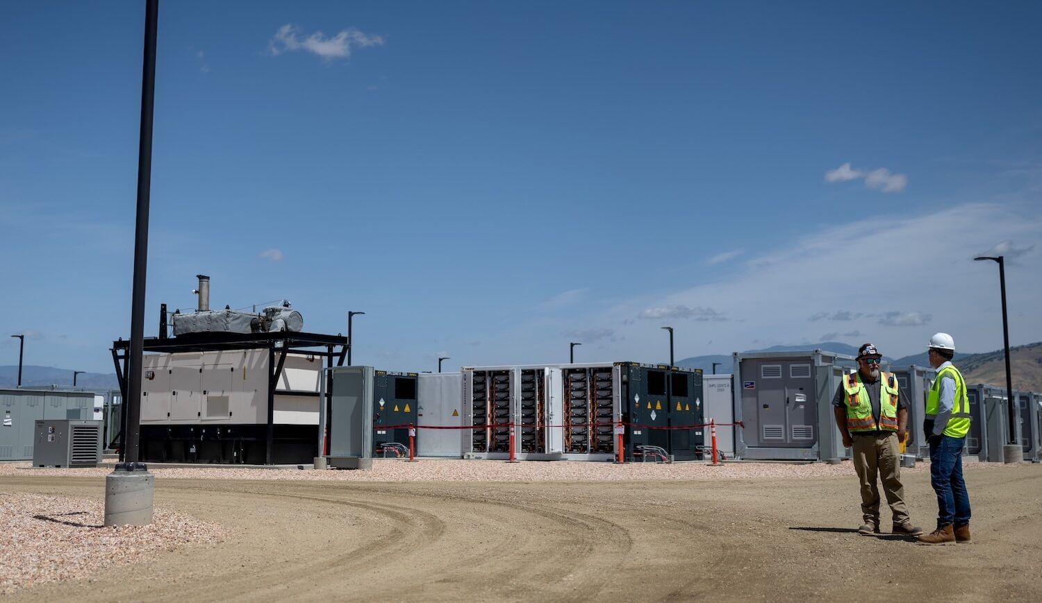 Two green-vested workers stand near batteries near a dirt road