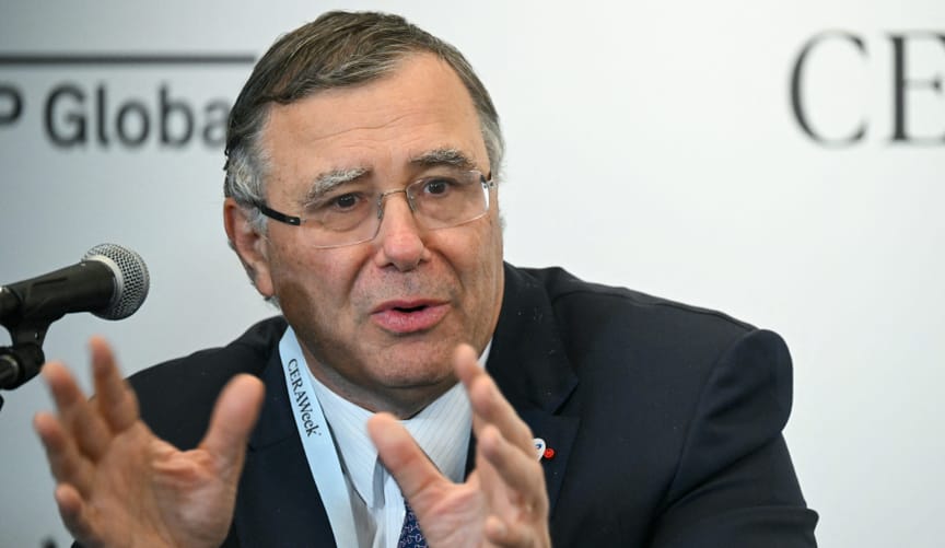 Man in a dark suit and light shirt with glasses speaks into a mic while seated