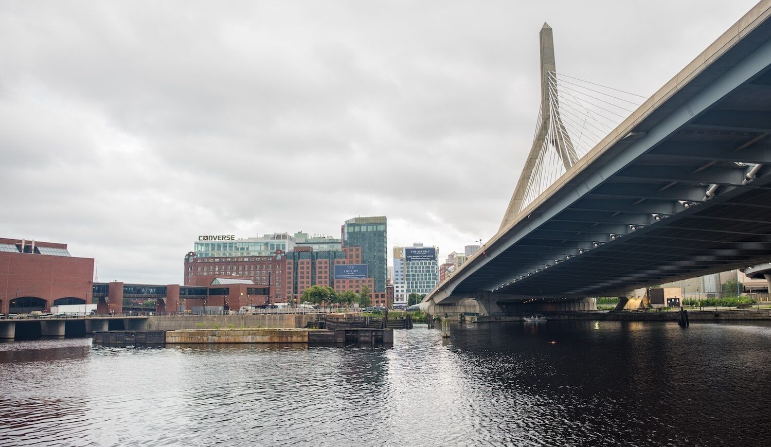 A section of dark river with brick buildings to the left and midrise buildings in the center, with a bridge to the right