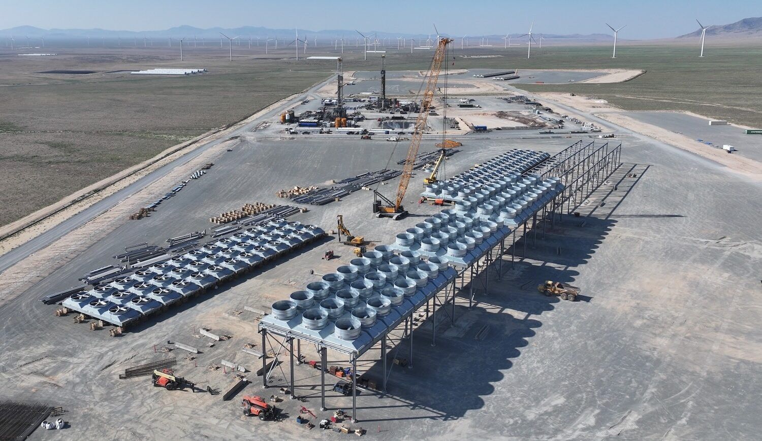 Geothermal power plant equipment on a long lot surrounded by a road with wind turbines in the distance