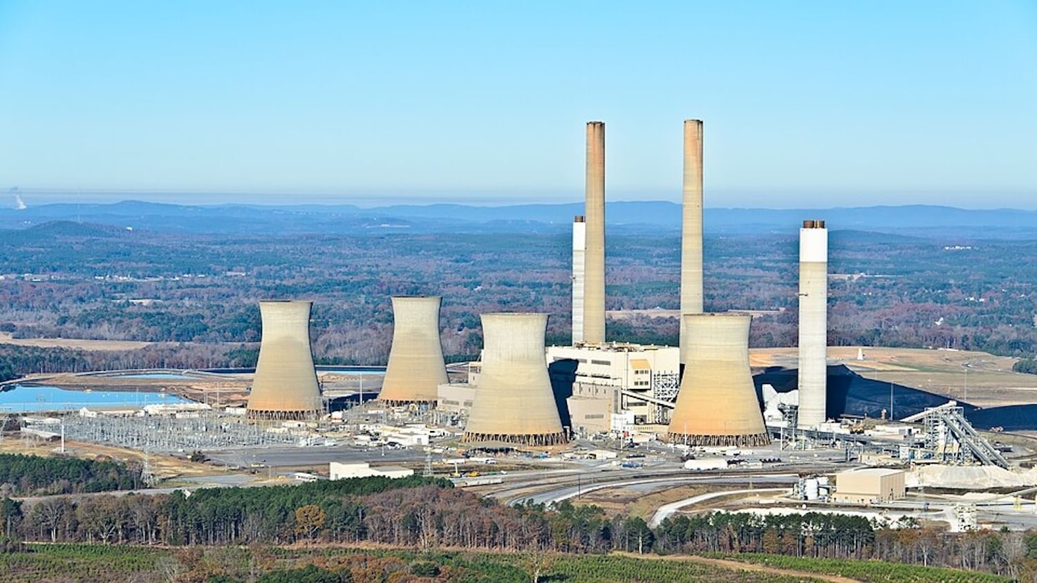 A large coal plant with multiple smokestacks is viewed from afar during the day