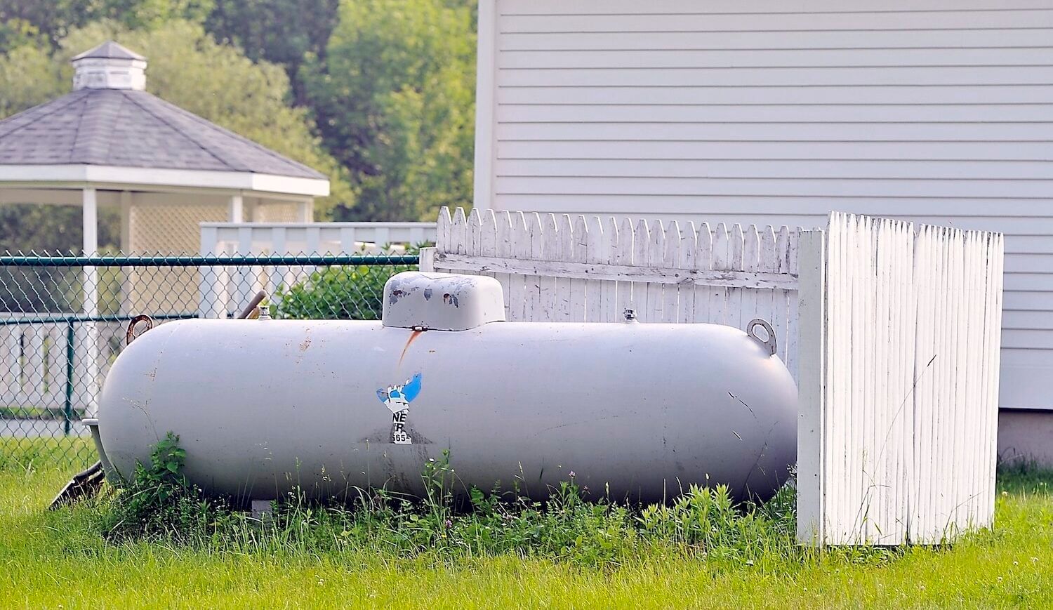 A gray torpedo-shaped propane heater on a green lawn shielded by a white picket fence next to a white building wall