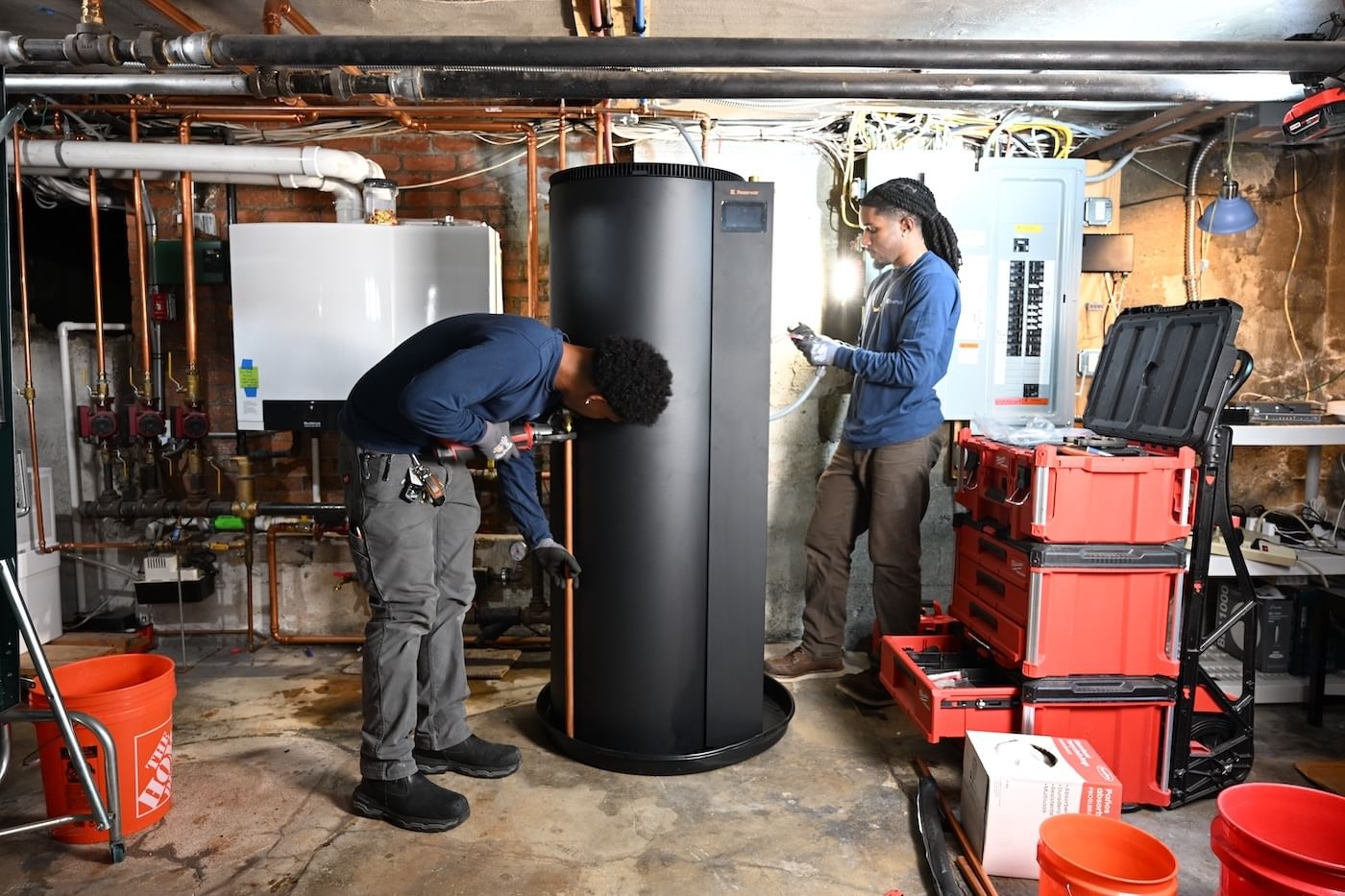 Two people in blue shirts and grey pants install a black water heater tank inside an unfinished utility space.