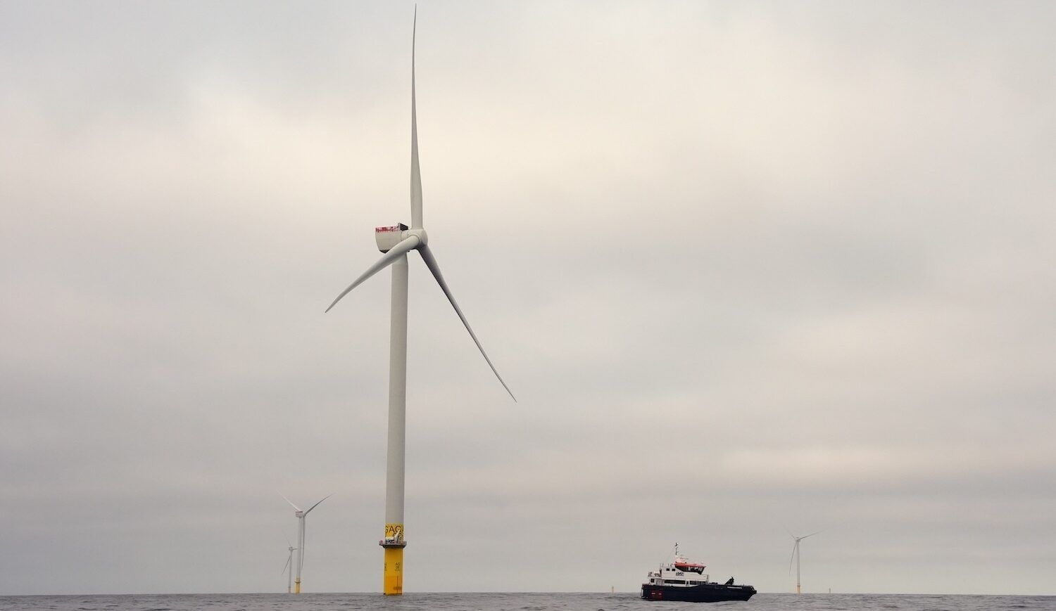 Wind turbines — one near center with a black-hulled boat nearby, and three others in the distance —  in gray ocean waters