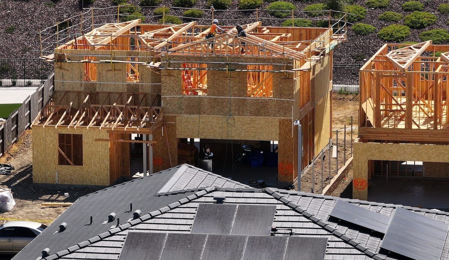 Houses under construction, with plywood and workers on roof; roof of house next door has solar panels