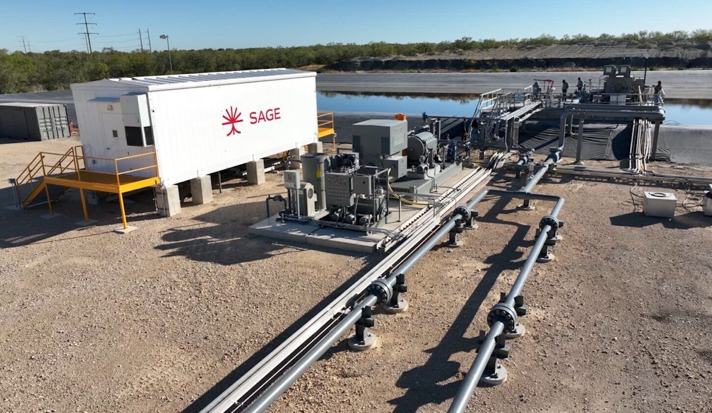Dirt lot with a white trailer that says "Sage" and various pipes and other equipment with a reservoir of some sort