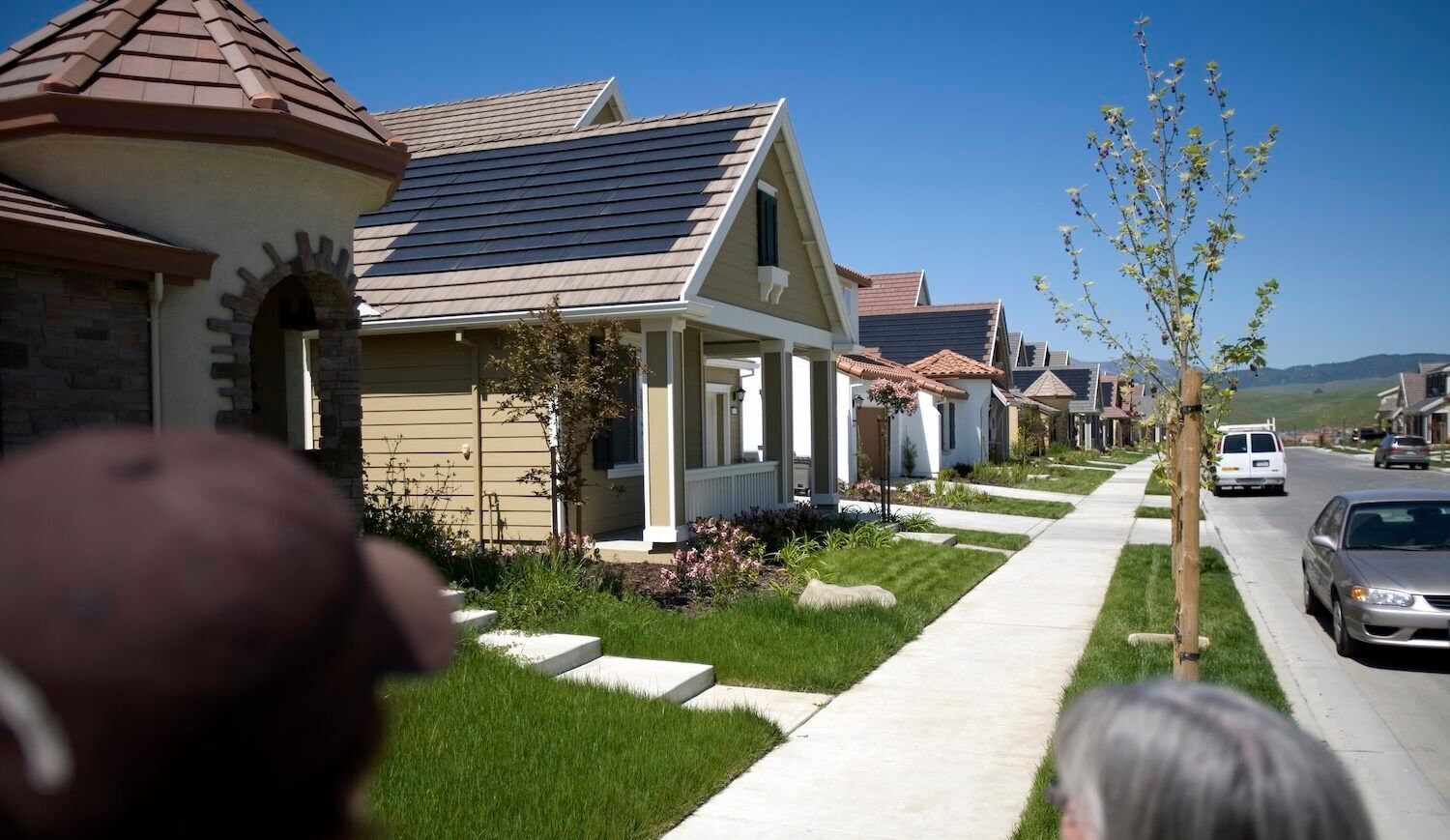 A row of two-story homes on the left, a sidewalk, and a street with a few cars; heads of two people on sidewalk in foreground