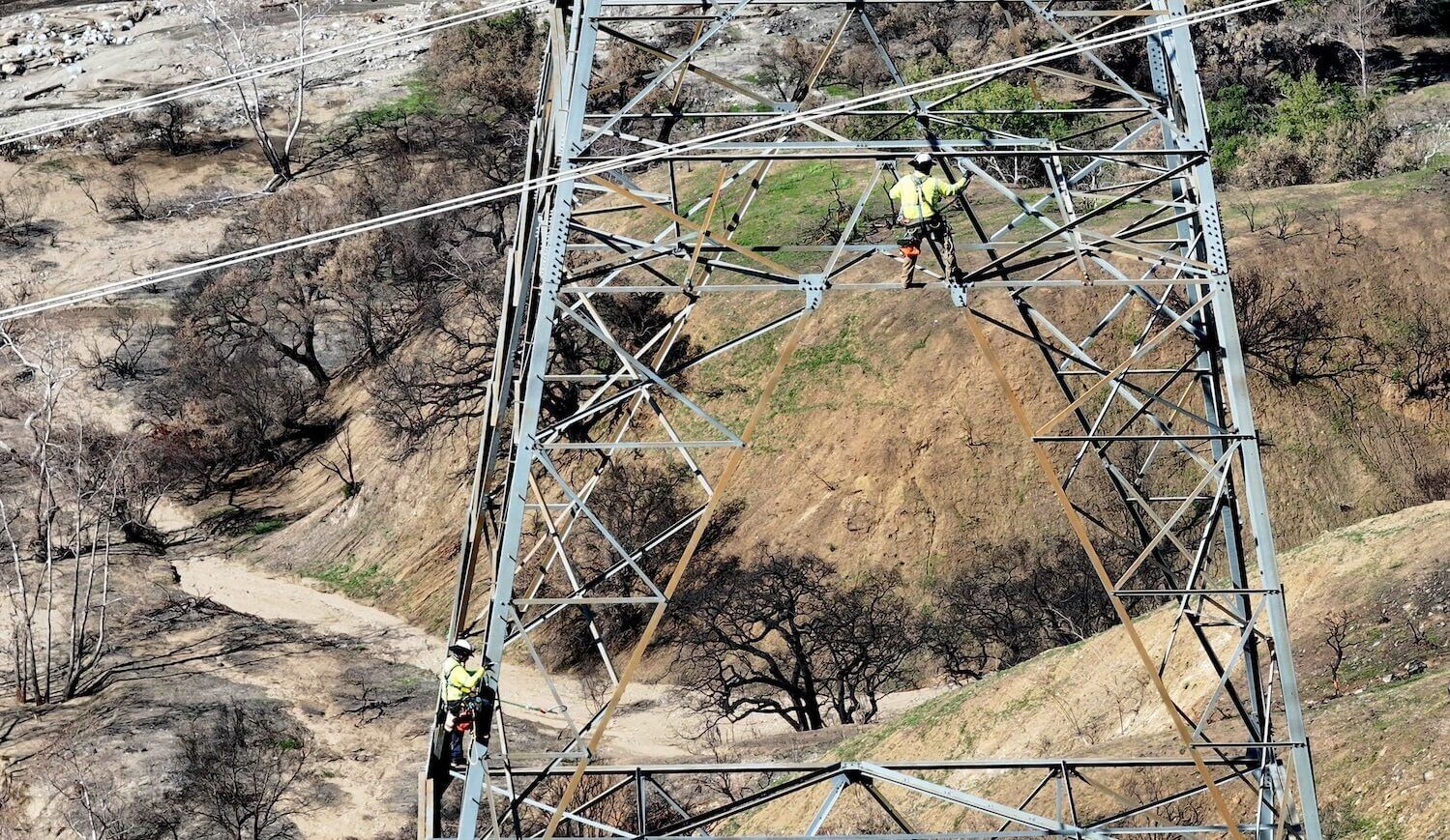 Two people in green vests on transmission tower with some charred landscape visible on the ground
