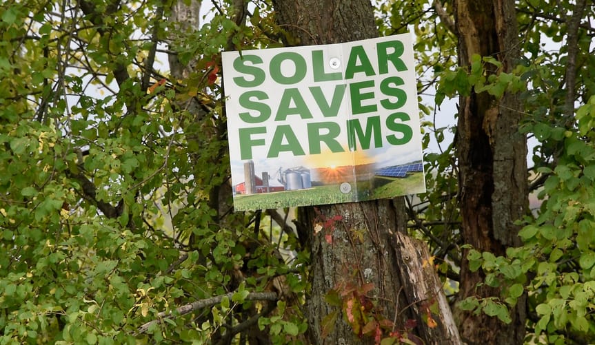 A ”Solar Saves Farms” sign, white with green lettering, on a tree