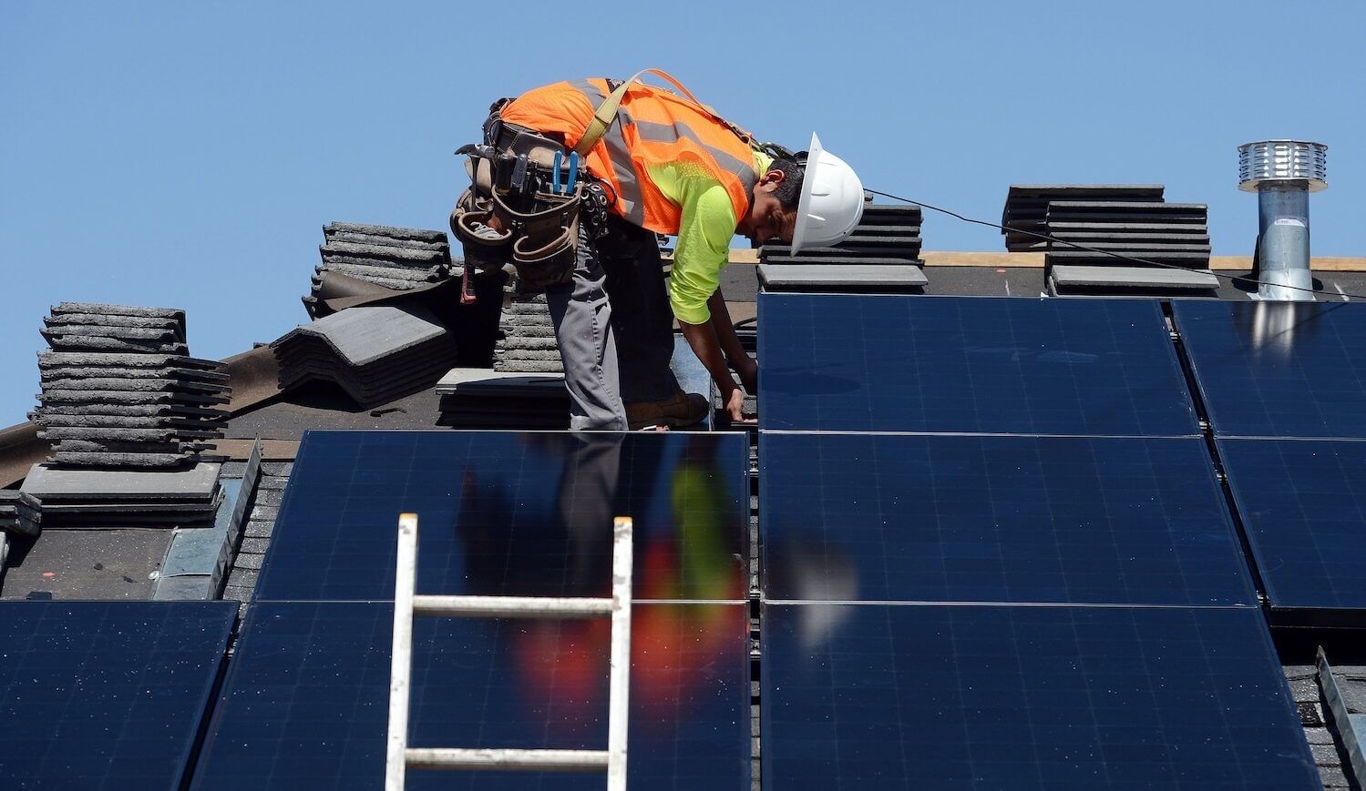 Worker in orange safety vest and white hard hat on roof installing solar panels; white ladder in front, roof shingles behind