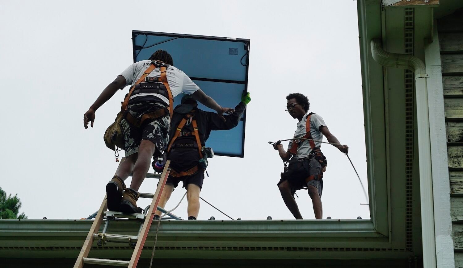Three workers on a roof—one holding a solar panel—seen from below.﻿