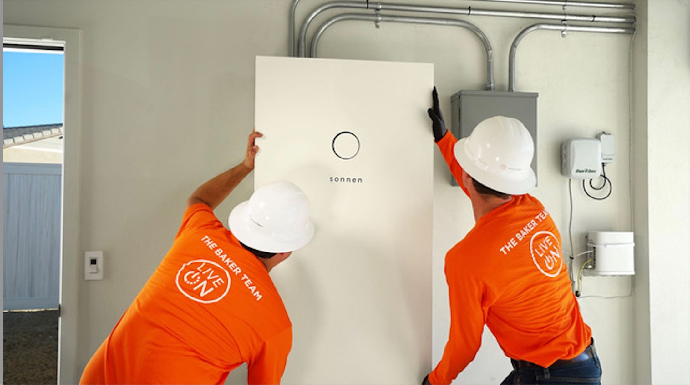 Two men in work uniforms and white hard hats install a metal rectangular white box in a home's garage