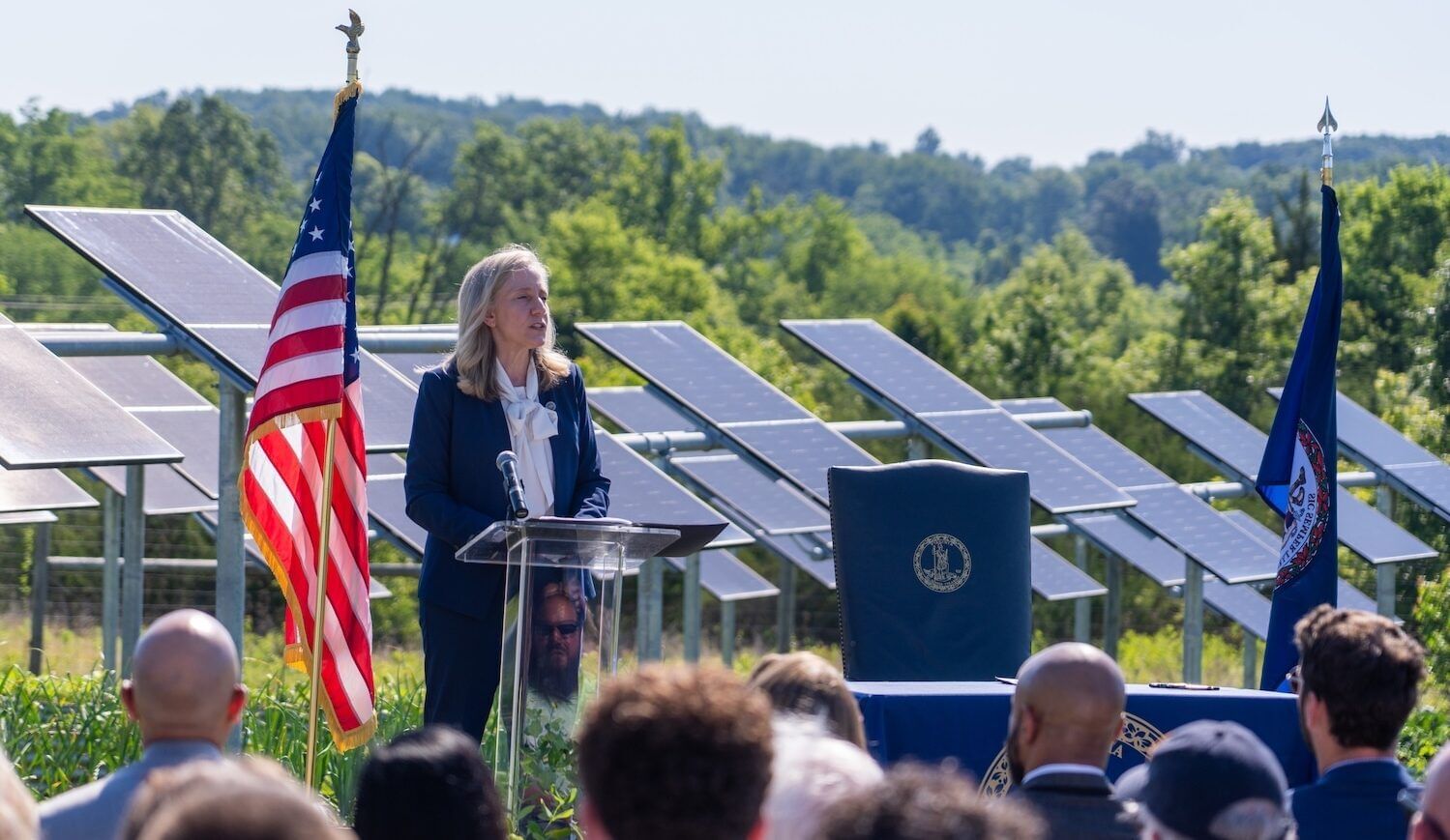 Governor in blue suit with white shirt speaks amid a field with solar panels to a seated crowd. American flag at her left.