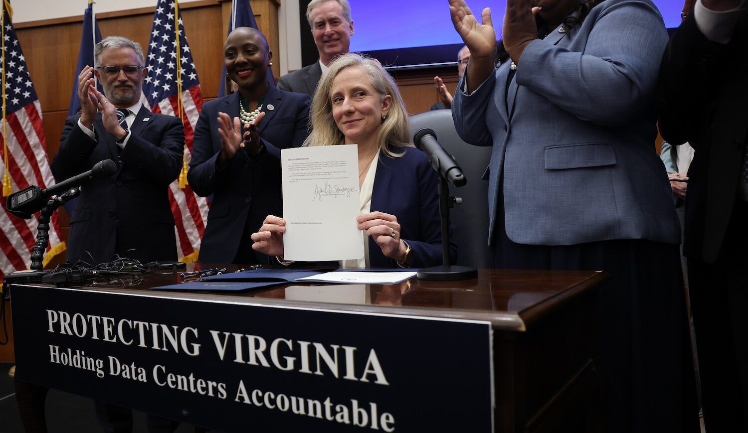 Woman sits at table showing signed order as people clap behind. Banner: Protecting Virginia: Holding data centers accountable