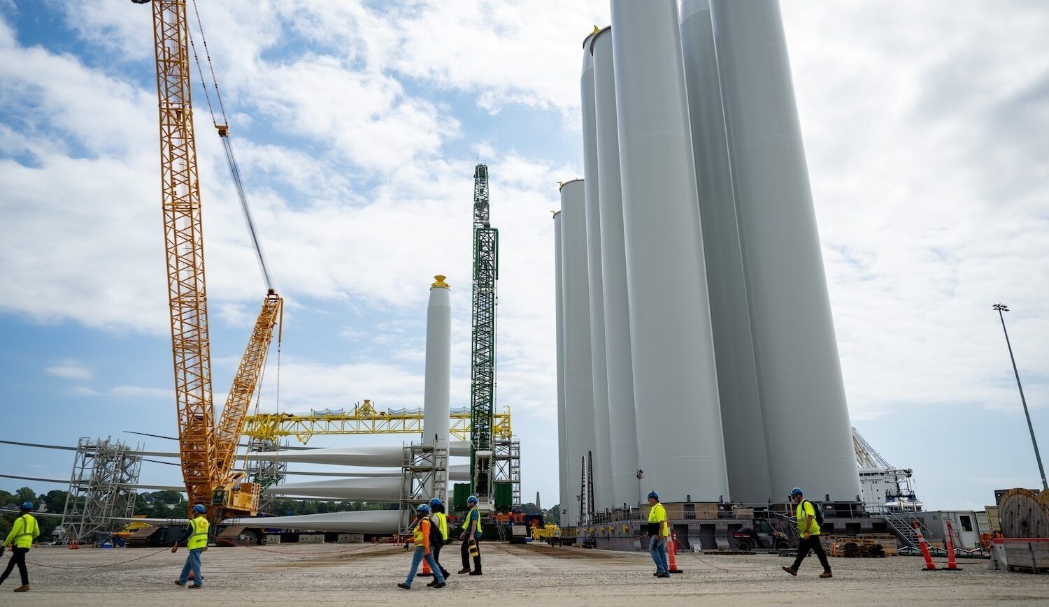Neon-vested workers walk past by turbine parts and cranes under a bright blue sky with some clouds