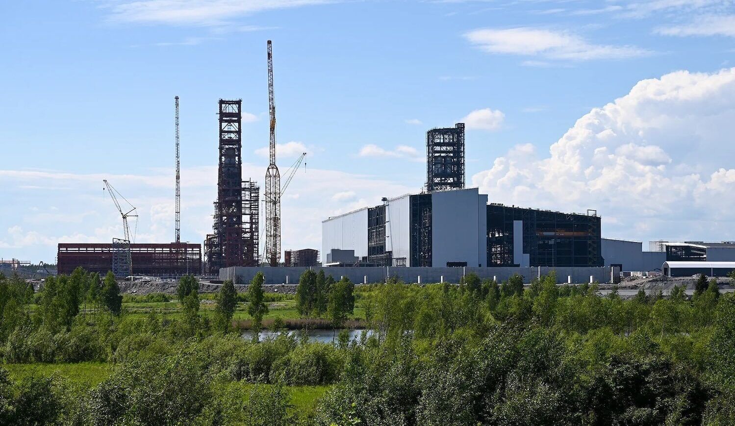 Steel plant with construction equipment; grass and small trees in front