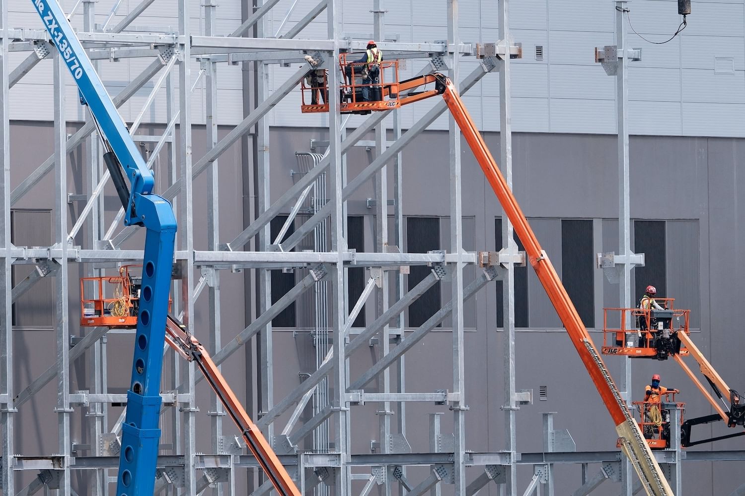 Construction workers on orange cherry pickers amid scaffolding near a data center wall being built