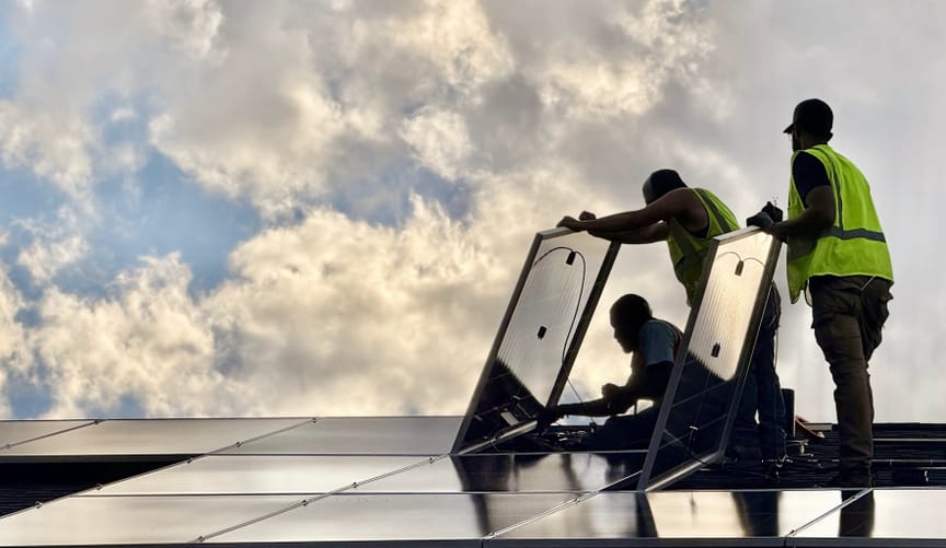 Three people on a roof installing solar panels with dramatic clouds overhead