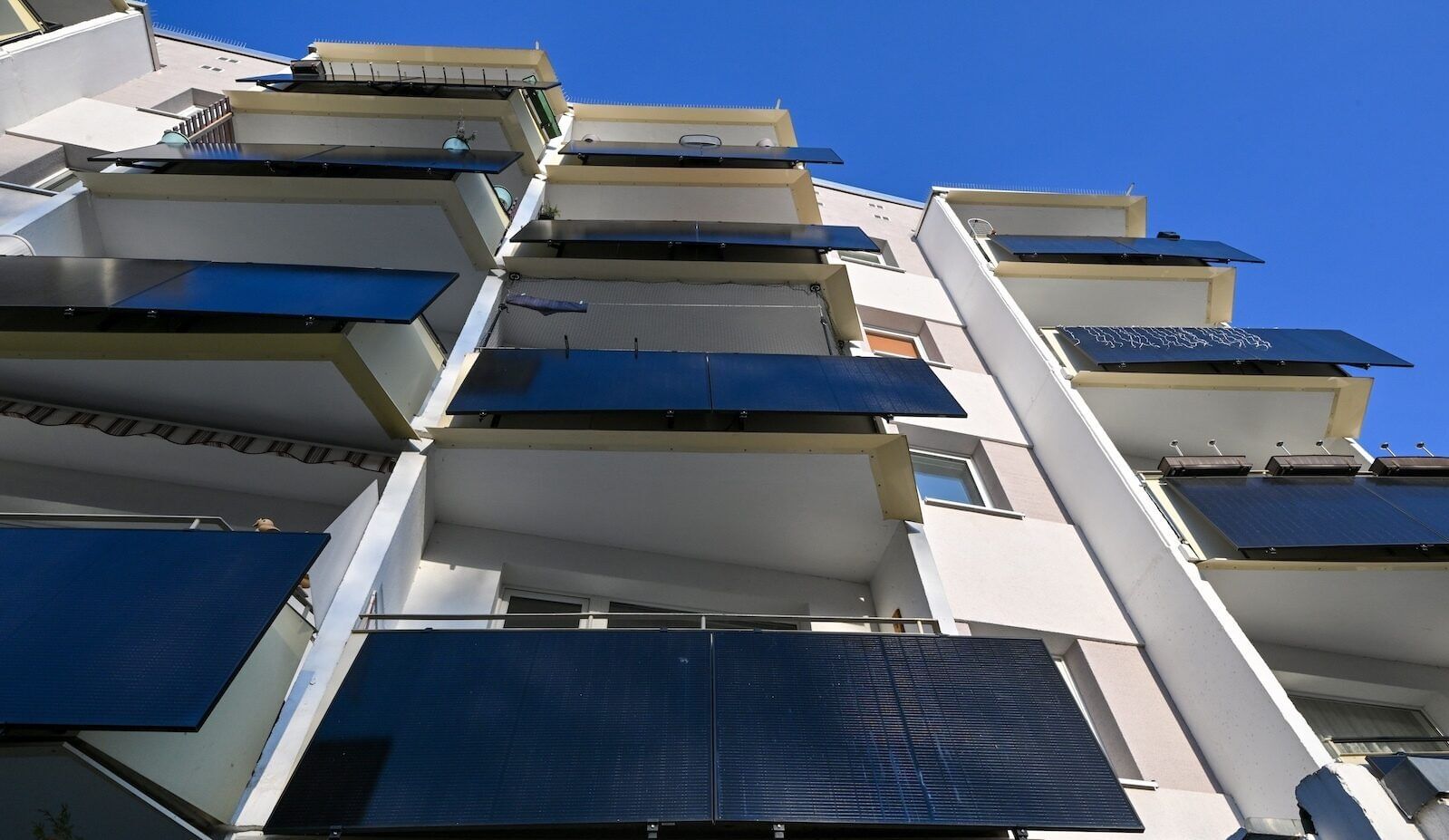 View from below of four stories of balconies with solar panels on a light-colored builiding