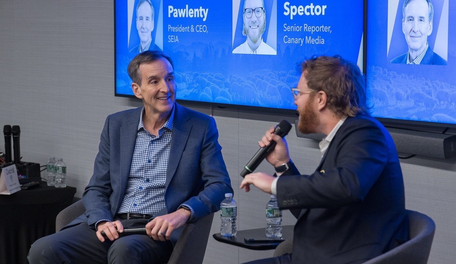 Two men sit in chairs, both in suits, one on the right holding microphone; background has names and images: Pawlenty, Spector