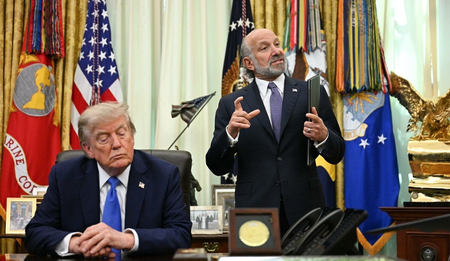 Two men, one sitting at a desk and one standing and gesturing, with flags behind them and curtains