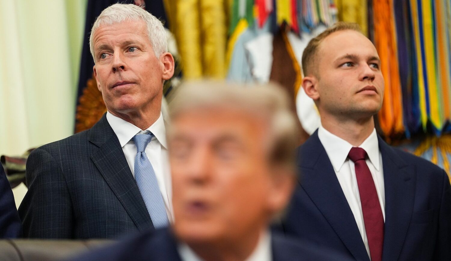 Three men in dark suits, one seated, with numerous flags behind them