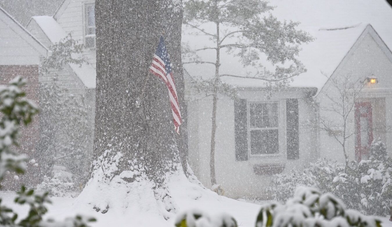 A white house with black shutters in a snowfall; a tree in the front yard has an American flag.