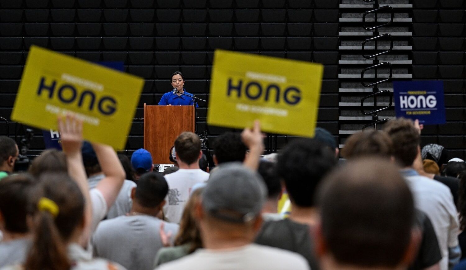 Woman behind a wooden lectern stands before a seated crowd; two yellow "Hong" signs are held aloft.