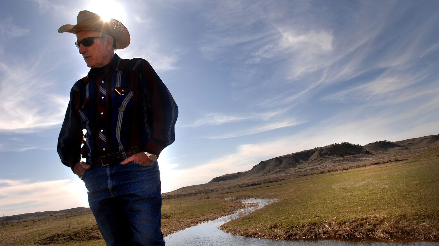 A man in a cowboy hat stands in front of a small creek on a vast plain