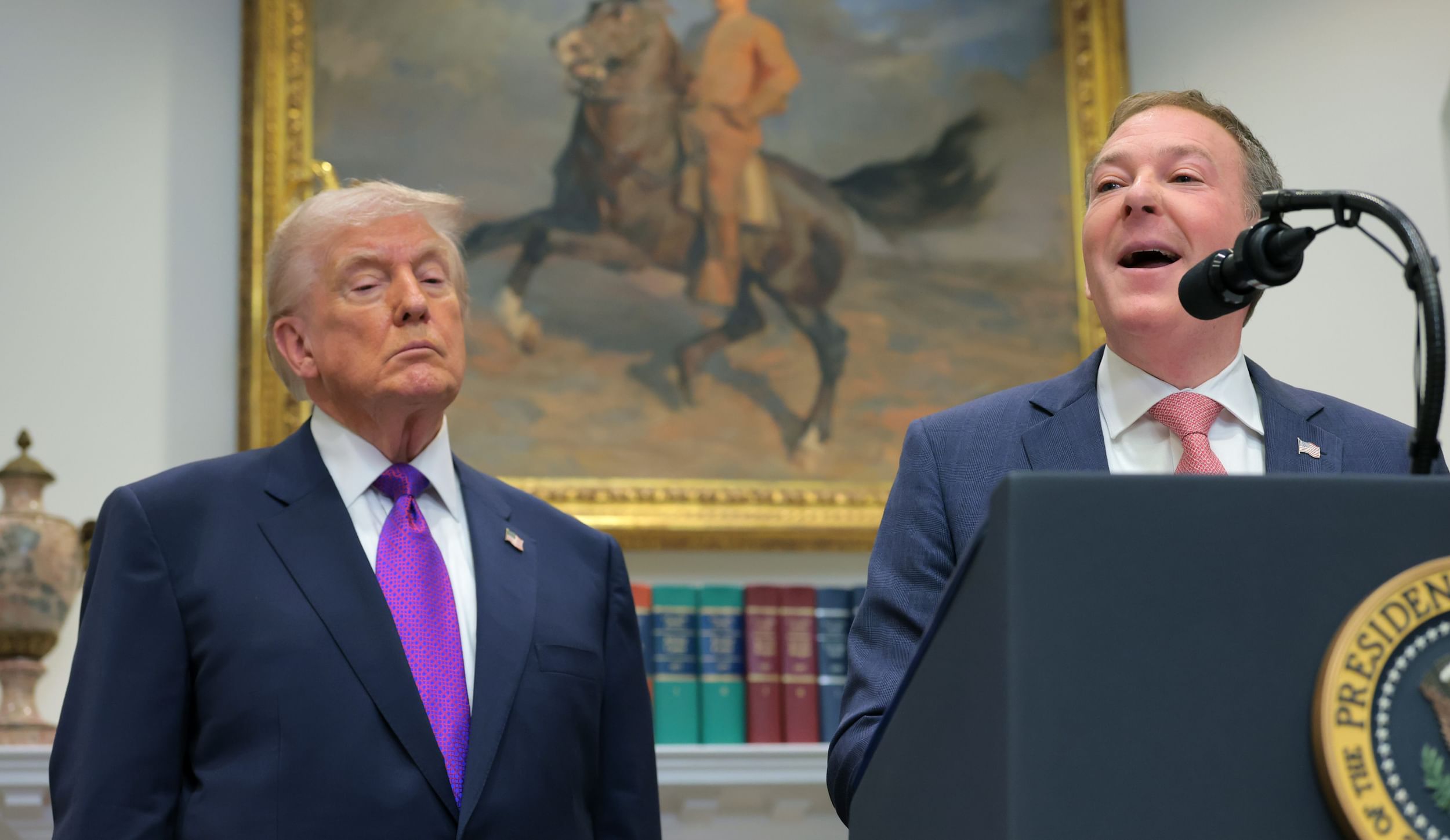 Two men in suits, one speaking at a lectern, with a painting of a man on a horse in a gold frame behind them.