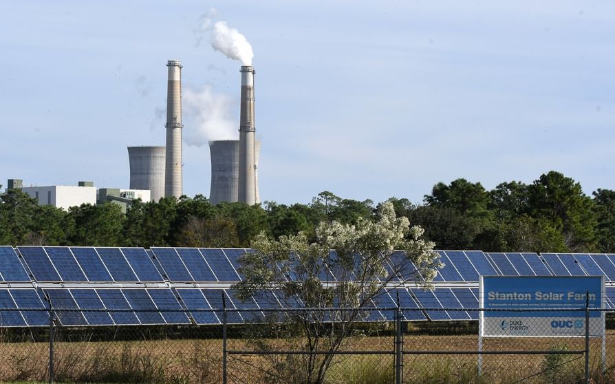 In the foreground is a large solar array. In the background are the stacks of a conventional fossil power plant