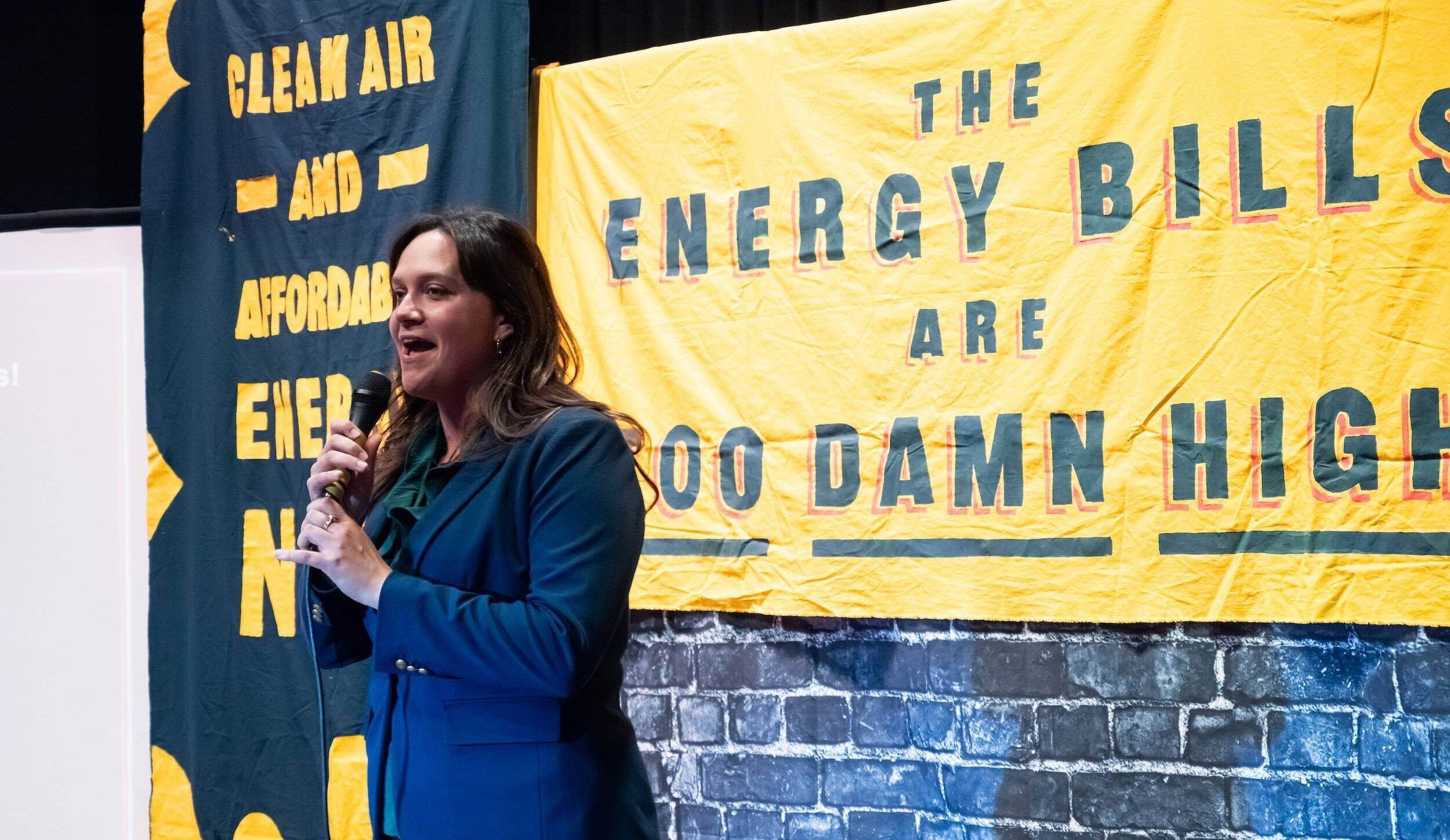 A person speaks into a microphone. Behind her is a sign that says "The energy bills are too damn high!"
