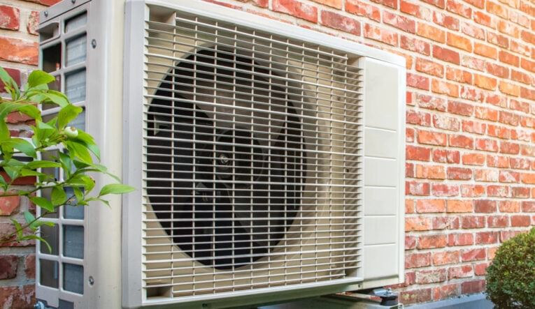 A rectangular beige heat pump installed on a red brick exterior wall of a home. Some foliage is visible around the appliance.